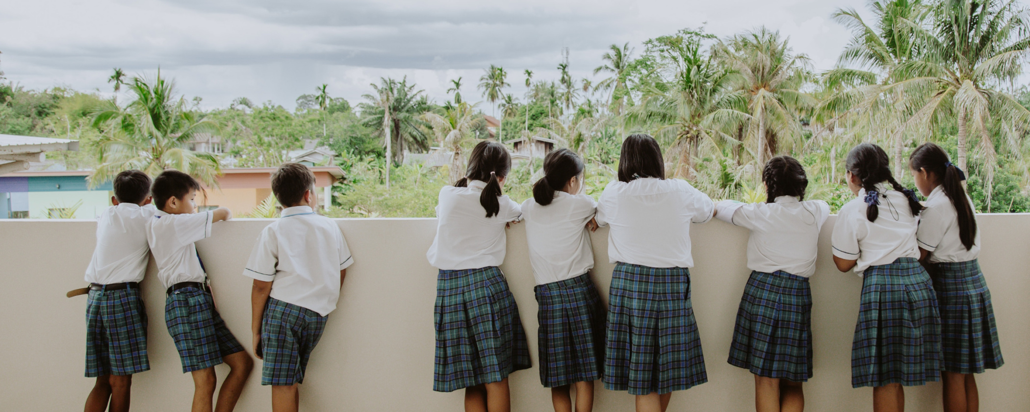 children dressed in school attire, with their backs to the photo, staring out from a balcony into a scenery with palm trees