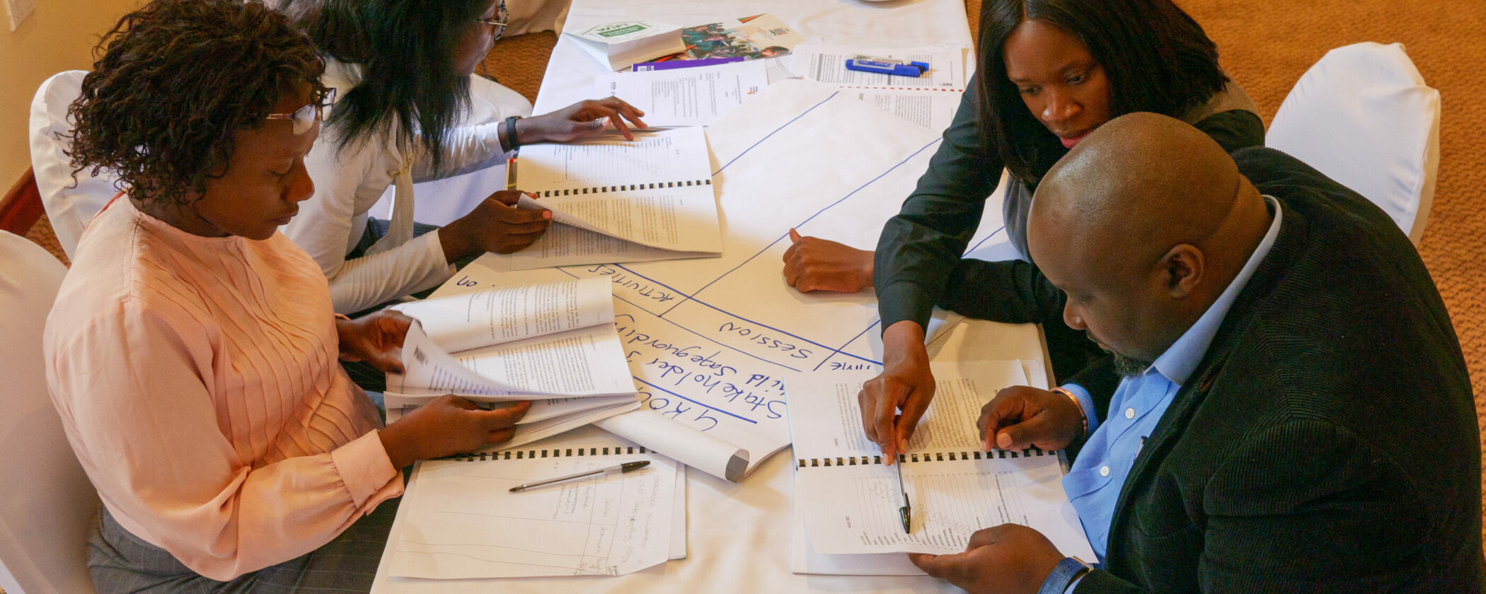 A group of people gathers around a table, checking notes during a working session on technical assistance to States.